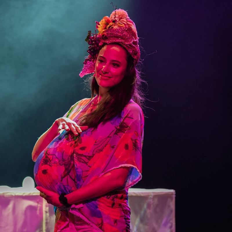 A women in a pink dress with a fruit hat on her head is smiling peacefully while holding what appears to be a pregnant belly, but is actually a balloon inside her dress. She is on a stage with colourful lighting in pinks, purples and bluees.
