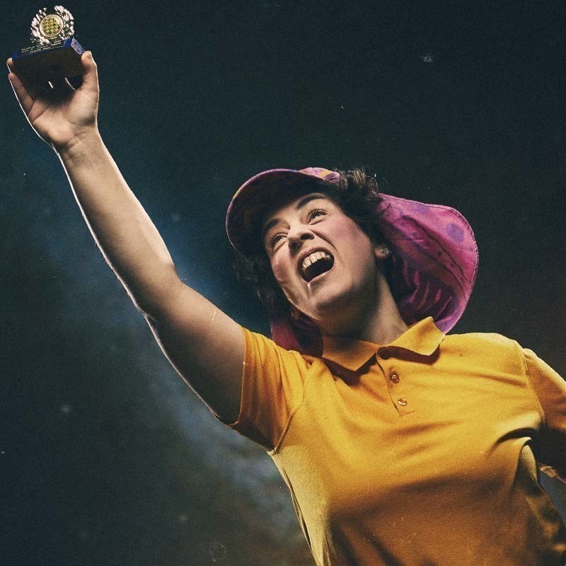 A comedian with brown curly hair wearing a pink visor and a yellow polo shirt holds a trophy up in the air, with a look of triumph on their face.