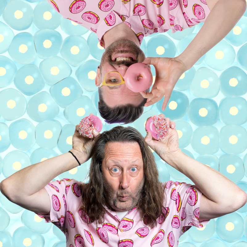 One man with long hair holds two pink iced doughnuts above his head like bear ears while an upside down man with yellow framed glasses holds one pink iced doughnut over one eye.