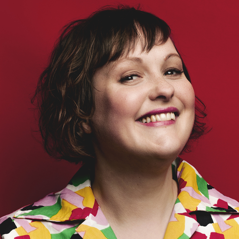 Josie is smiling looking to the camera with a cheeky grin of the image wearing a colourful top with multi coloured triangles on it. She has short brown hair and red lipstick on, and is against a red background.