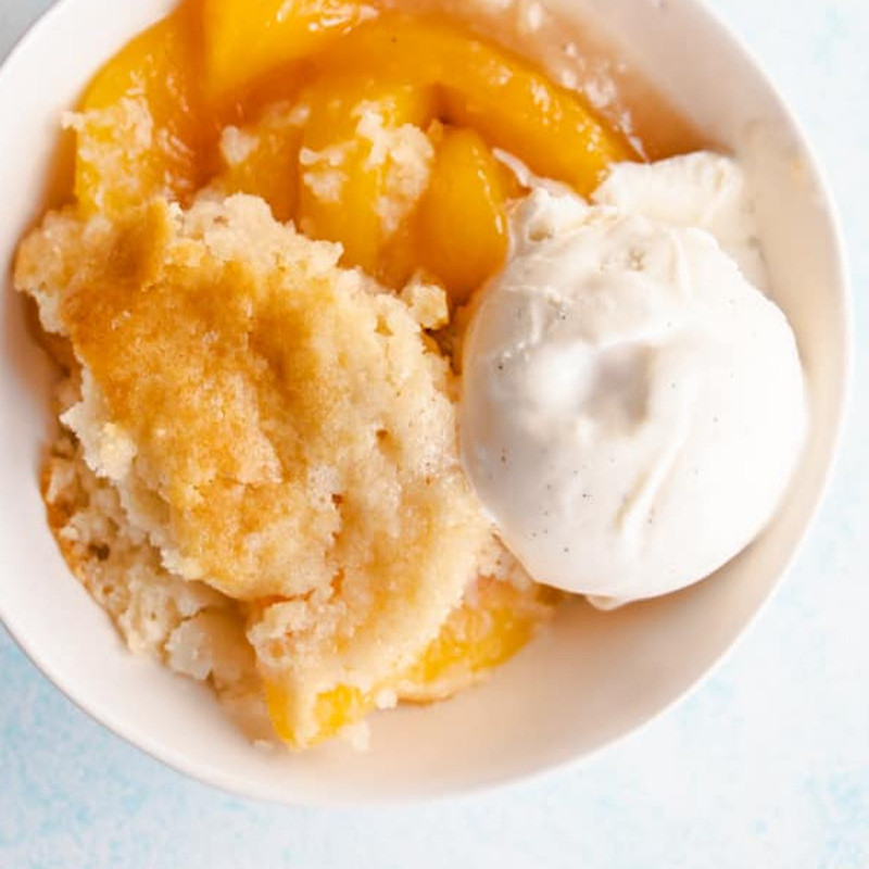 A close up image of white bowl with orange peaches crushed biscuit with one scoop of white ice-cream.