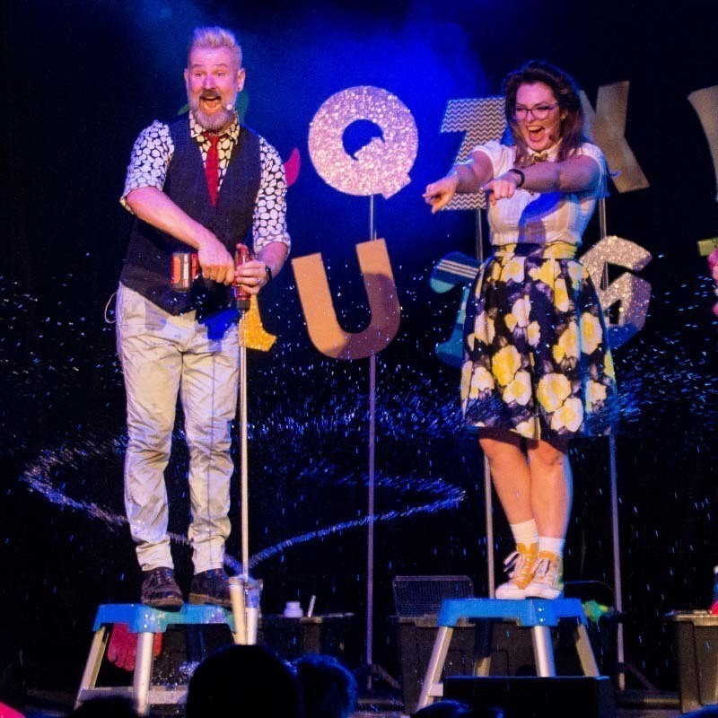 A man with silver hair and a grey beard is holding a power drill attached to a series of pipes that are spinning in a bucket and flinging water out in two jets. A woman with long brown hair is laughing and pointing into the audience with two hands. The man and woman are standing on stools in front of a colourful collection of large letters.