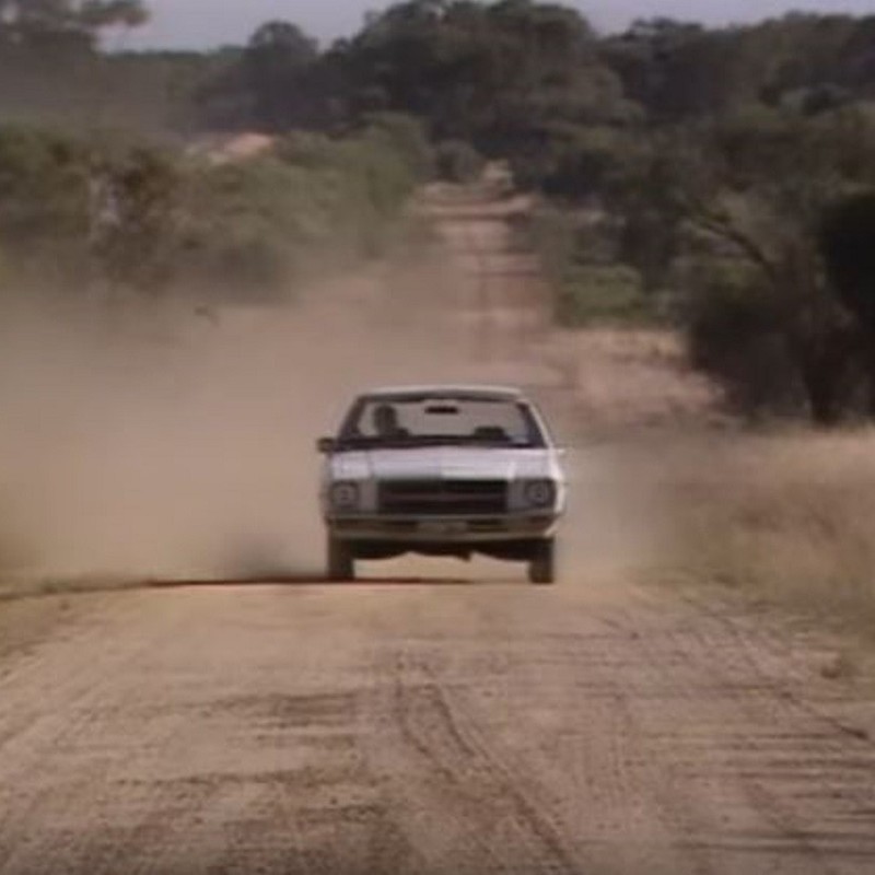 Wide Open Road - tribute to the Triffids - A white car is driving down a dusty road in the middle of the outback. The image looks like a still from an 80's movie.