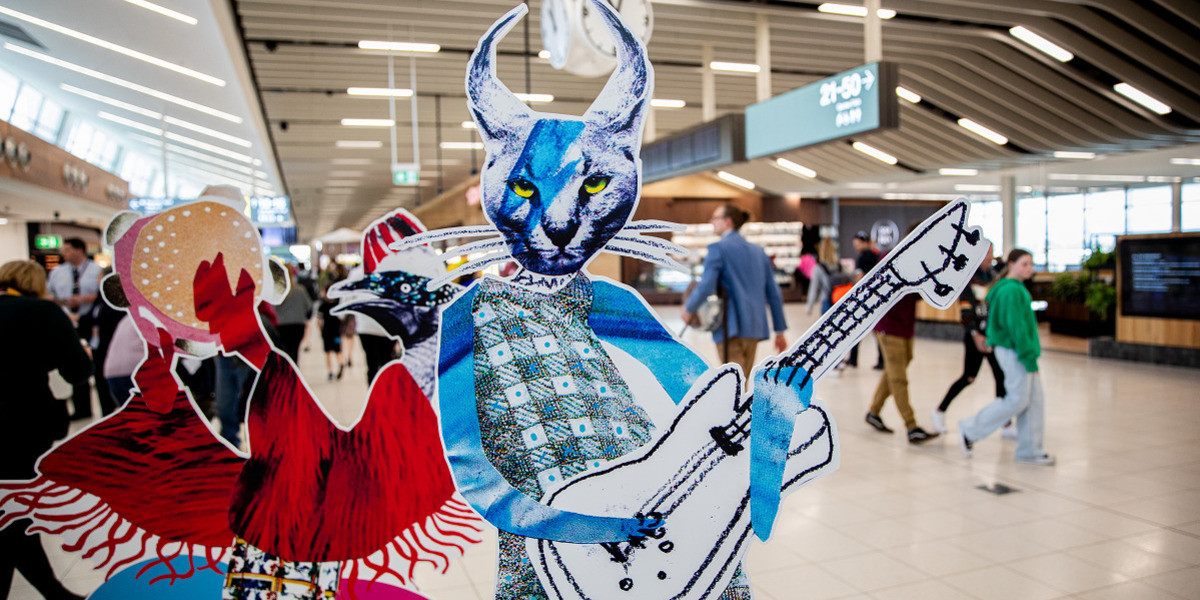 'Menagerie of Characters' Installation - Two characters from the 2024 Poster artwork are free-standing in the foreground of the terminal with a crowd and tv screens in the background. The characters are a giant cat playing the electric guitar and an cassowary playing the tamborine.