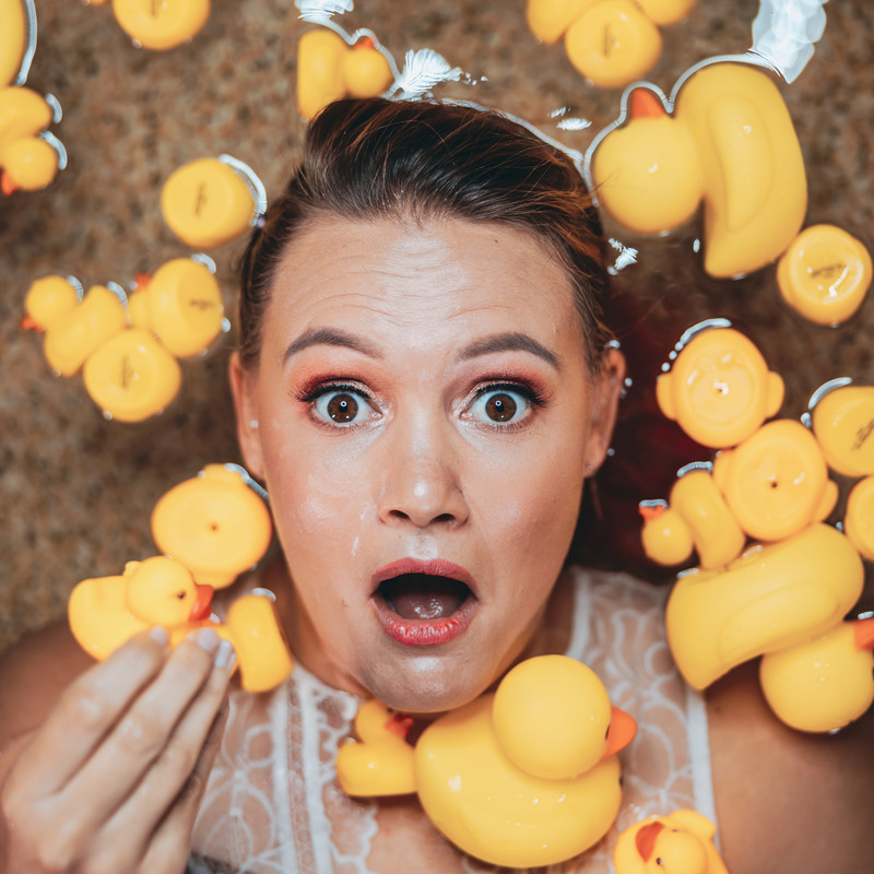 A woman with brown hair and fair skin is lying in a shallow pool of water surrounded by yellow rubber ducks. She has a surprised/shocked expression on her face, she wears a white lace top.