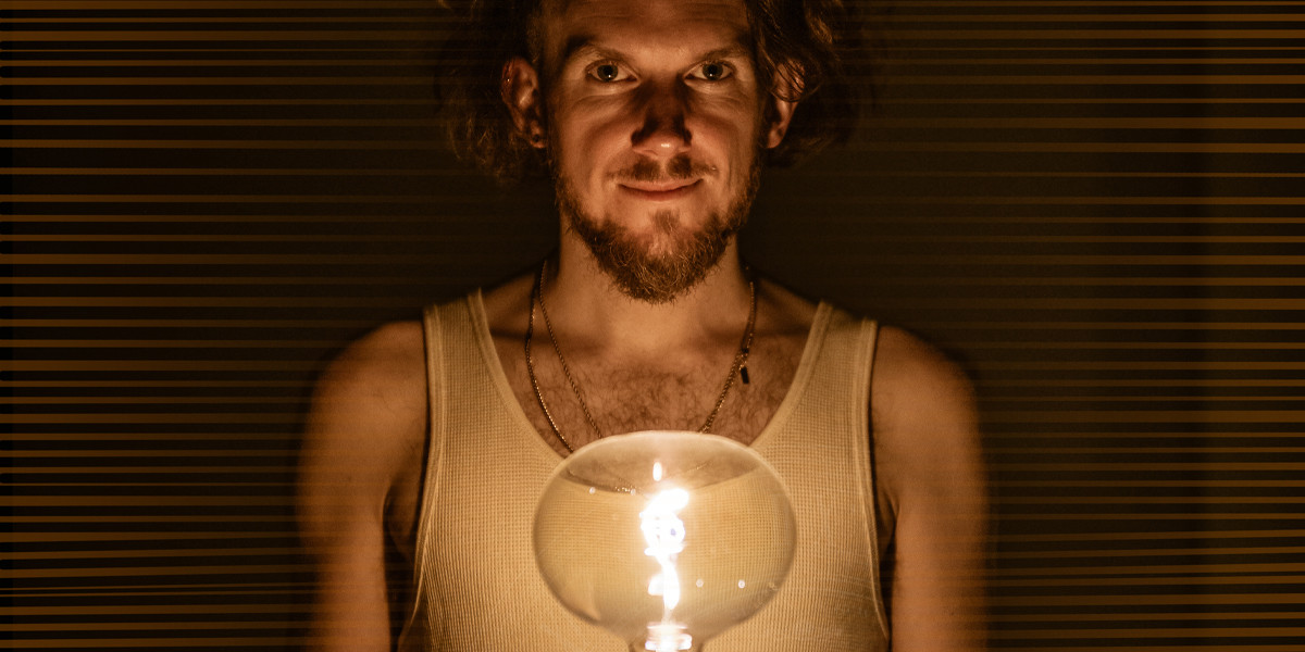 Helios - A man in a white vest stands lit by a warm bulb in front of him, looking directly into the camera lens.