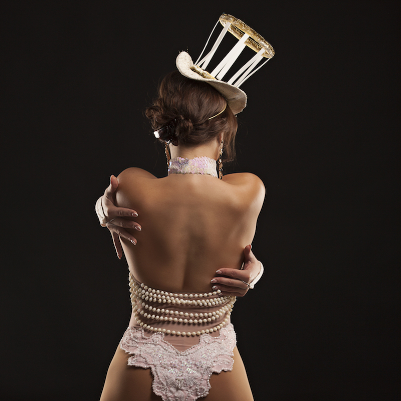 Woman stands with her back to the camera with her arms wrapped around herself. She is wearing a pink and black top hat with pearls, and underwear in pink lace with strings of pearls across her lower back.