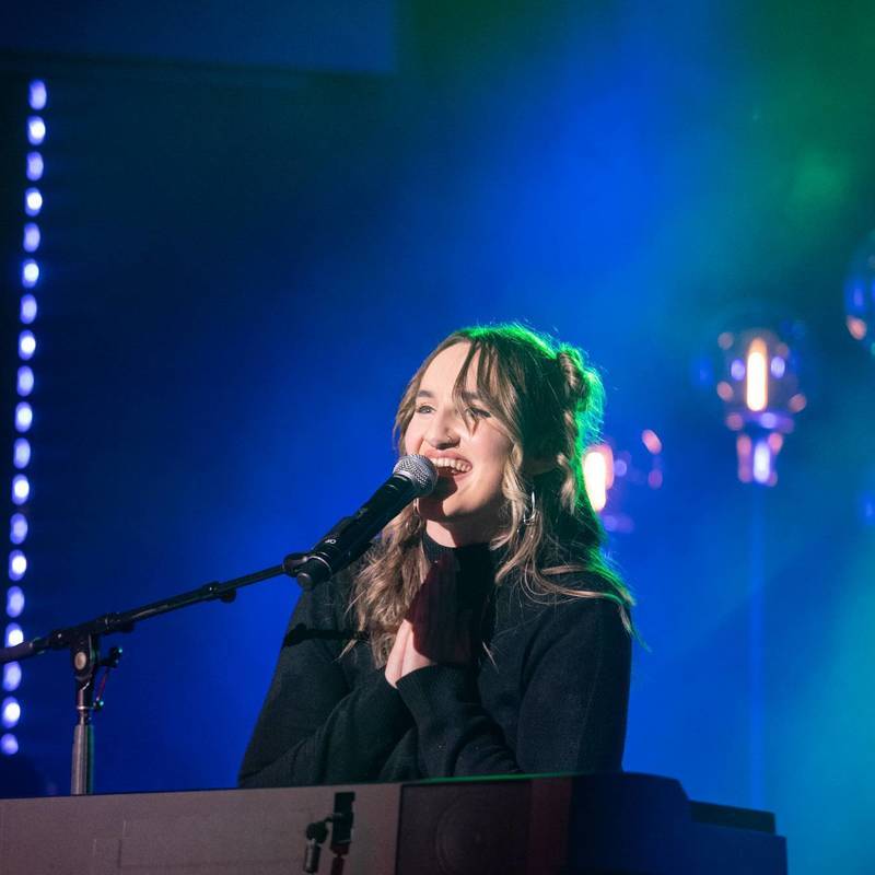 Image of a young woman seated at a piano singing into a microphone with stage blue stage lighting behind her.