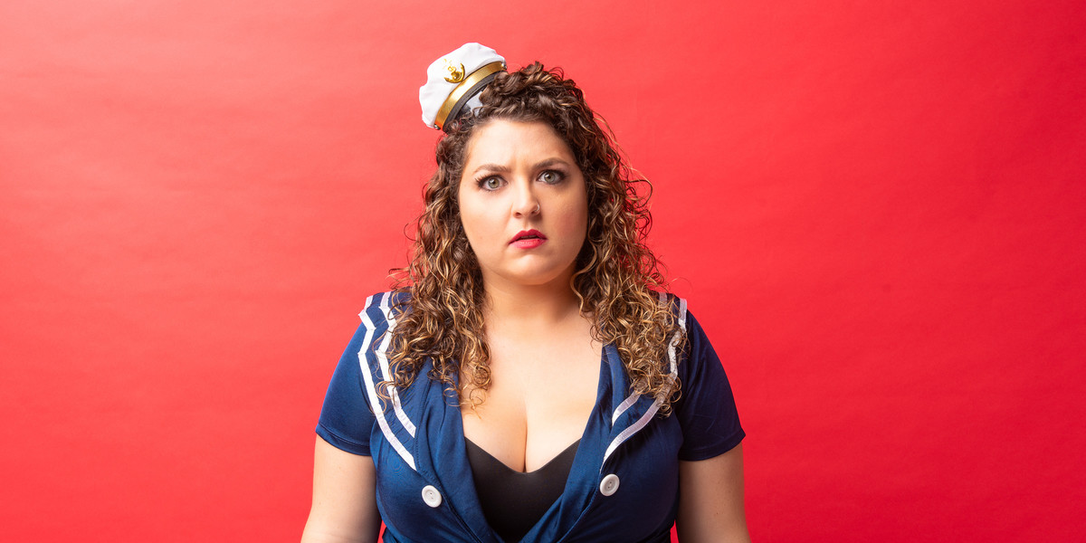 A woman with curly brown hair and a tiny captain's hat stares into the camera in front of a red background