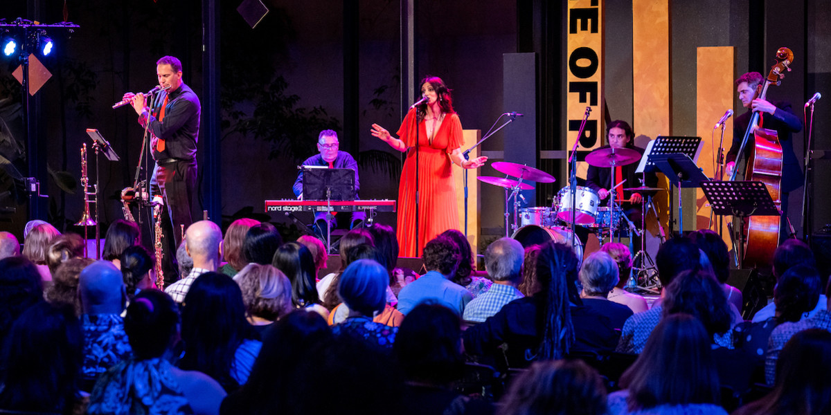 A whole jazz band performing on stage, with a female vocalist in a red dress and male band members playing flute, keys, double bass, and drums. The audience seems to listen very carefully.
A singer is singing on stage with her band, their hands are in the air. There are two dancers on stage as well, their hands are raised in the air as well. The all crowd is standing and dancing along with the music. 

Buona Sera Signorina - State Theatre Centre of WA - Photo  by Sean Breadsell.