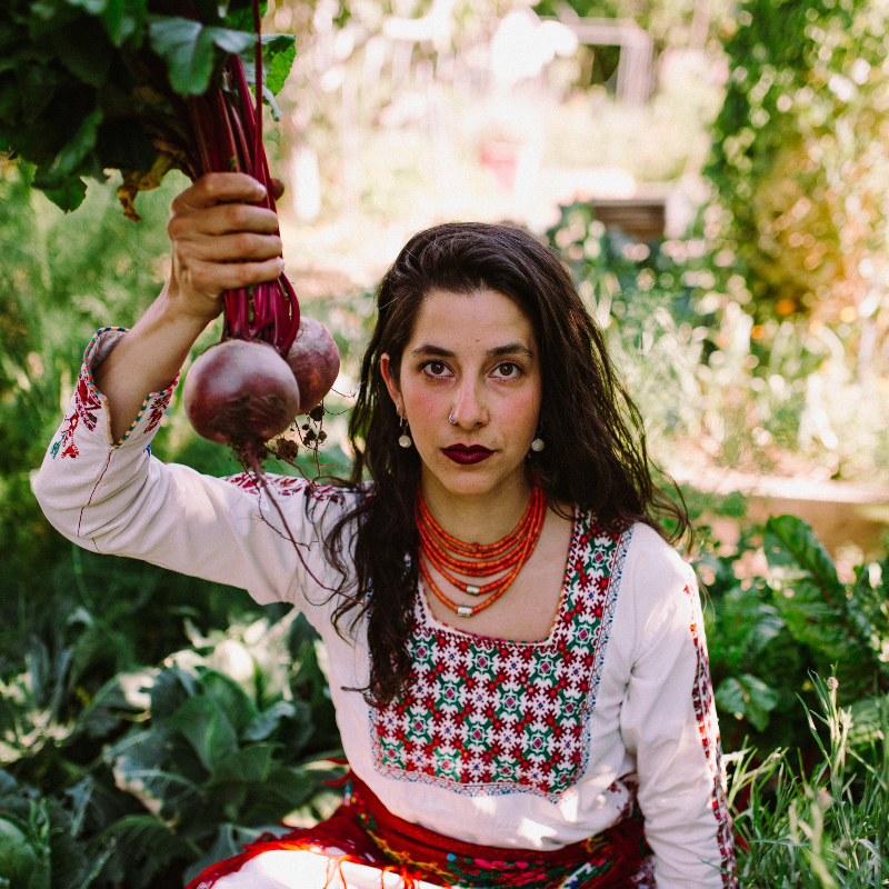 A woman with dark hair, white earings, white and red dress and red beaded neckless kneels holding a bunched uprooted beetroot in the garden.