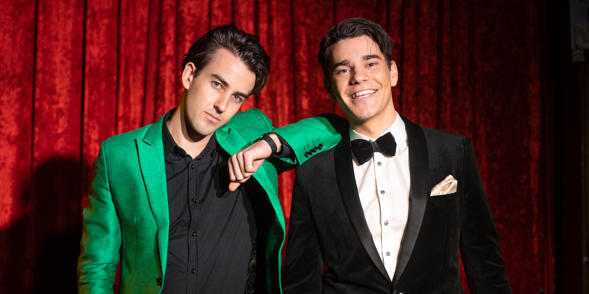 Two men in dapper suits in front of a red velvet curtain