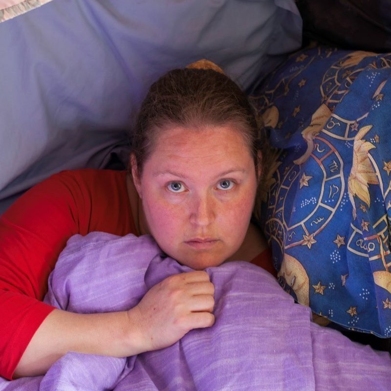 Sheltered - Young woman looking directly at the camera curled up in a nest of blankets.
