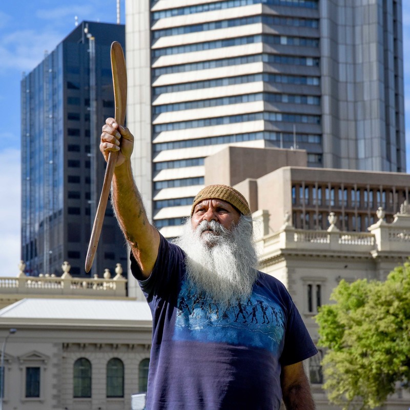 Kaurna Elder, Major Sumner, holds up boomerang to sky and looks out over Victoria Square. He is wearing a beanie and has a long grey beard.