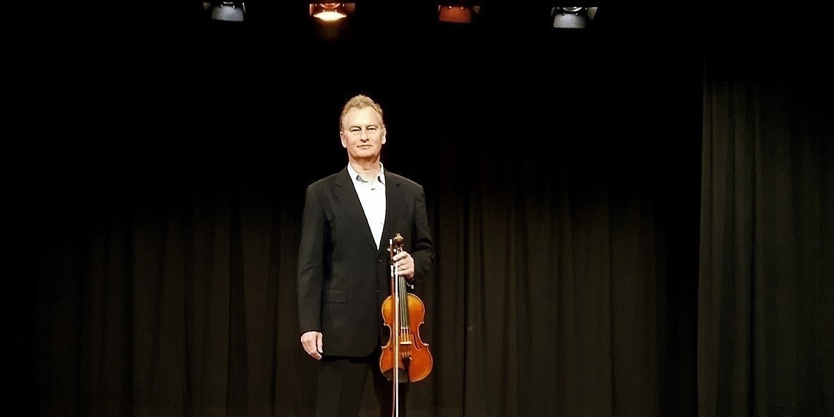 CANCELLED - Jonathon Glonek: Solo Violin - Jonathaon Glonek stands 3/4 profile against a white background, looking towards the camera in a black suit and white and blue grid check collared shirt. His violin is tucked under his arm.