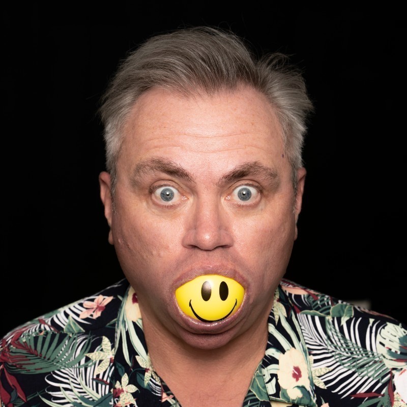 Groove Terminator's Right Here, Right Now: A Celebration of Fatboy Slim - A head and shoulders shot of a man with grey hair, wearing a tropical print shirt and facing the camera. In his mouth he holds a yellow ball with a black smiley face on it.