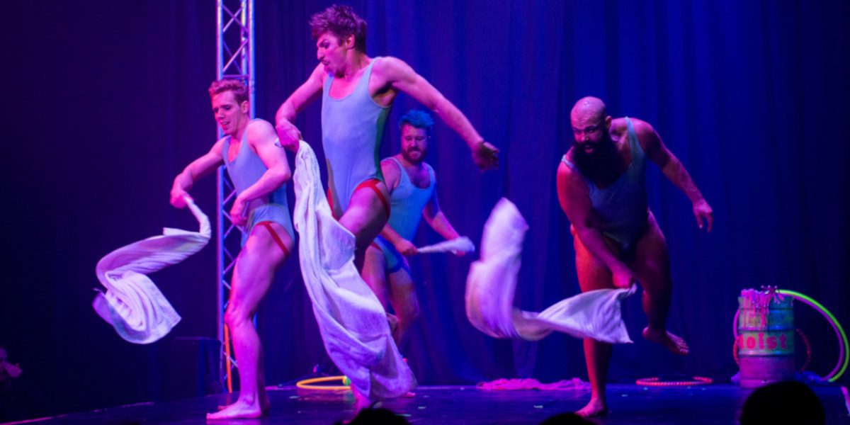 4 men running around a stage in various states of distress and excitement. one man in the back is winding up their towel to hit the other men on their bums. the background of the photo is dark. and there is a soft blue light on the performers.