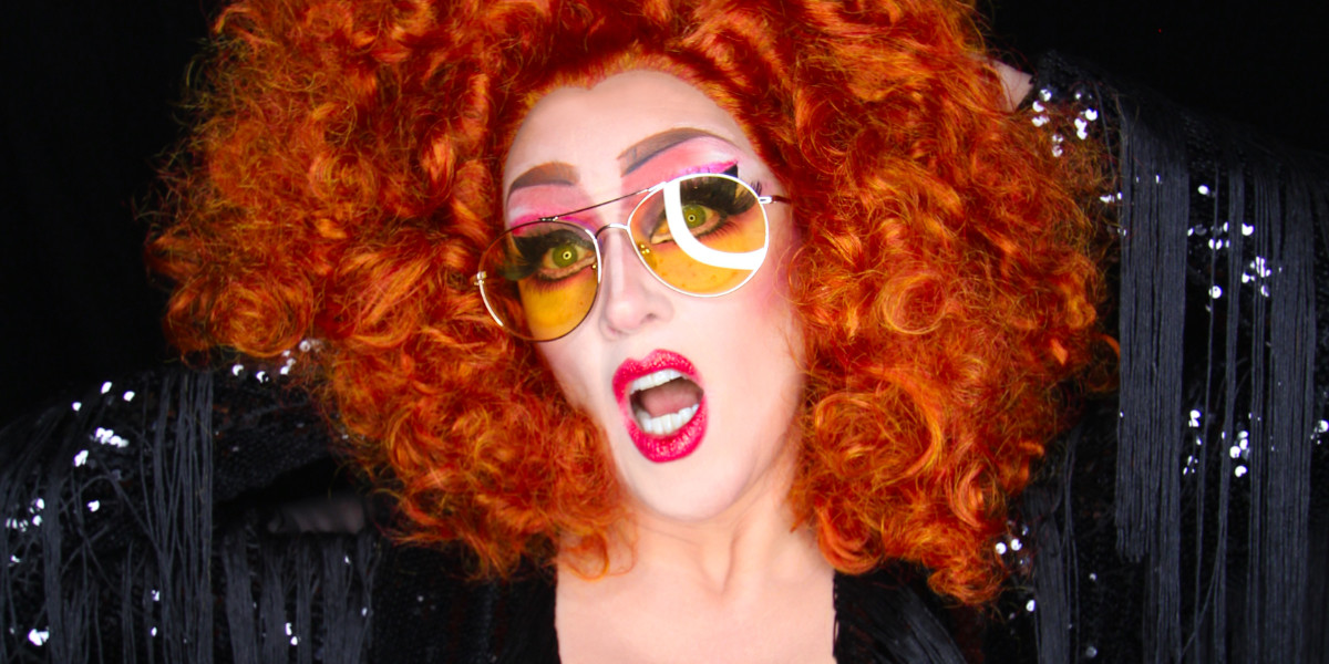 Beautiful middle-aged porcelain-skinned cis woman wearing black sequins and yellow-tinted glasses, her hands behind her mass of curly, red hair, lips parted as if in a drag war cry.