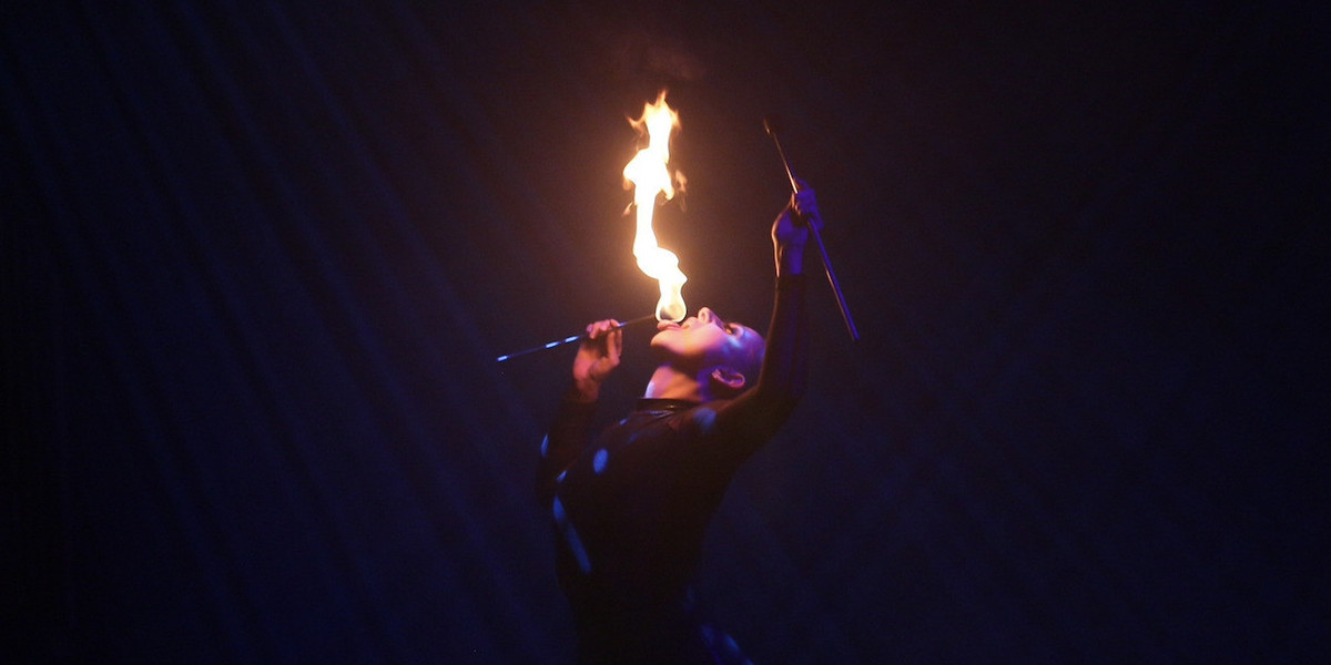 A fire eater holds a burning fire wand to her tongue as the other arm is extended up into the air holding another extinguished fire wand.