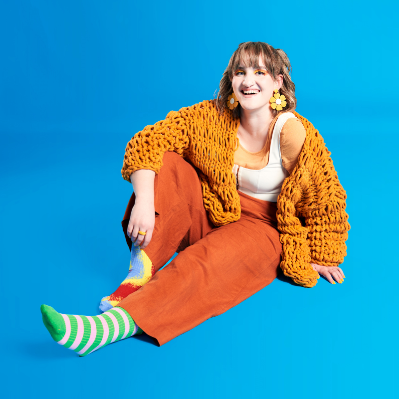 A woman is sitting in a relaxed position dressed in vibrant orange clothes with flower earrings. She is wearing two very different socks one multicoloured and the other is striped. The background is a bright blue