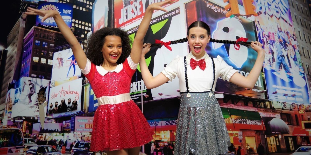 Annie in a bright red dress and Matilda in a school uniform stand in front of Times Square.