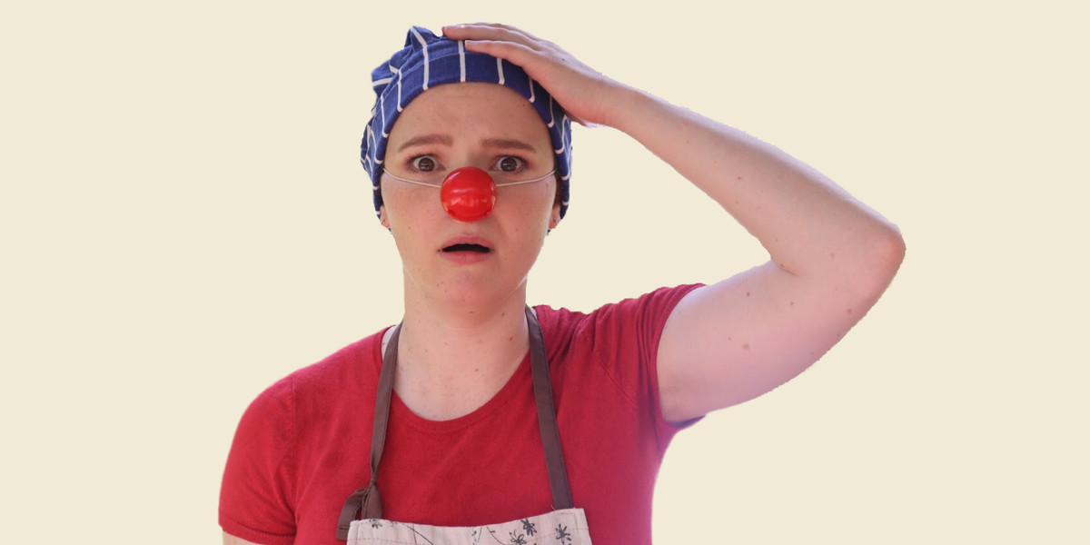 A clown faces the camera with her left arm raised up to clasp her head, while the other arm is at her side. She is wearing a red short-sleeved shirt, a small blue chef's hat with white stripes and a brown and white apron with a brown apron string, as well as a red nose. She has a worried/regretful expression and open mouth. It is a mid shot with an off-white background.