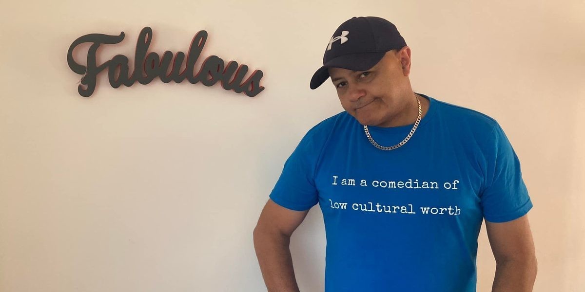 British comedian Nik Coppin stands by a 'Fabulous' sign with a blue T-shirt which reads, 'I'm a comedian of low, cultural worth'. A joke from a review he received at a show, which was printed for him by the reviewer.