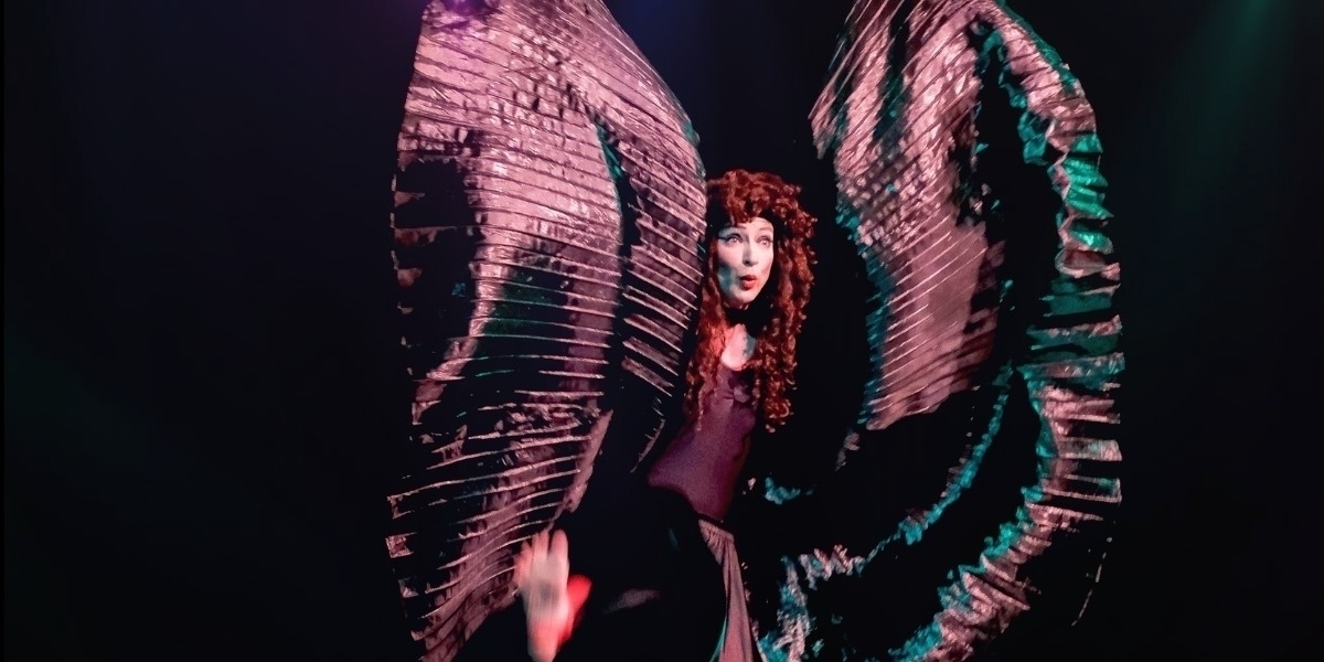 Sarah-Louise Young is dressed in a metallic jumpsuit with matching pleated material "wings", she's flailing these wings to imitate a butterfly. She wears a curly red wig (in the style of Kate Bush), a puckered-lipped look of surprise, and kicking her foot to the side.