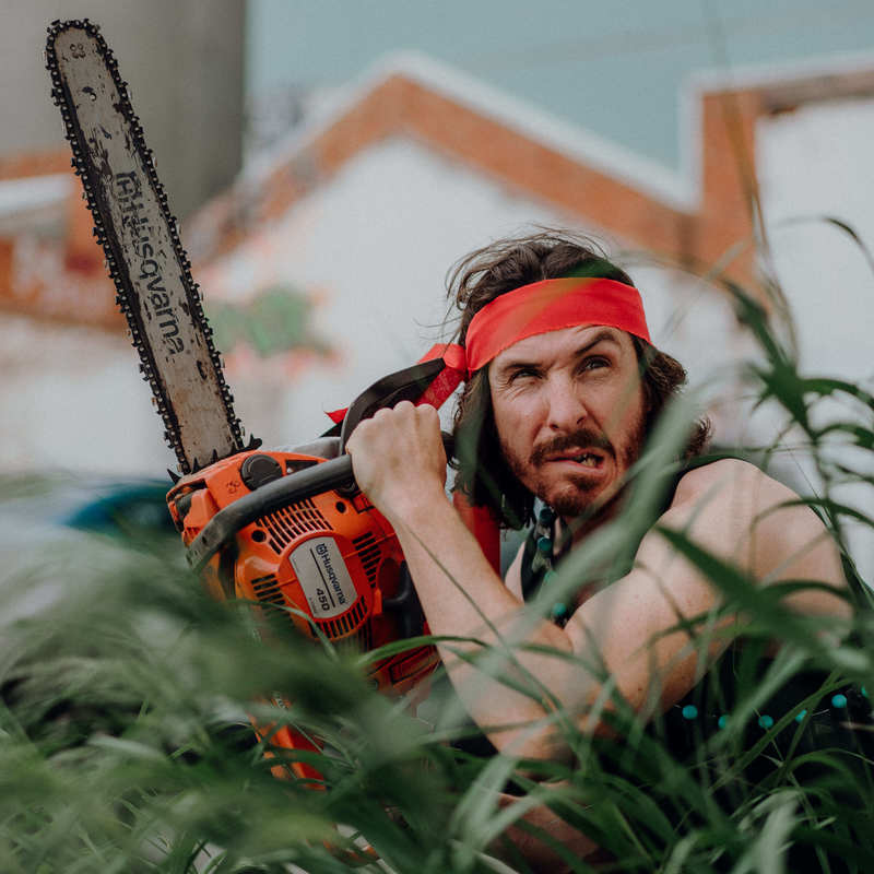 Man with a red headband on ducking down in long grass, holding a chainsaw.