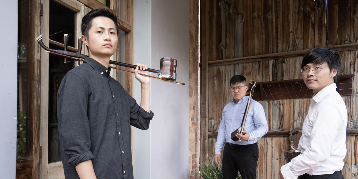 Three adult male of Chinese descent each holding Chinese fiddles, standing in an alcove, each in a staggered position and varied pose.