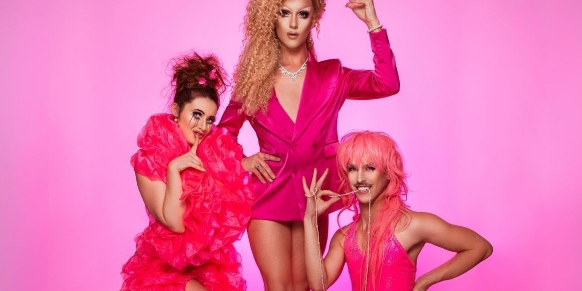 HUSH - Female with large pink boa and bare feet stands next to a drag queen in a pink suit jacket and no pants. Kneeling on the ground is a drag artist in a long hot pink dress. They are all in front of a pink backdrop.