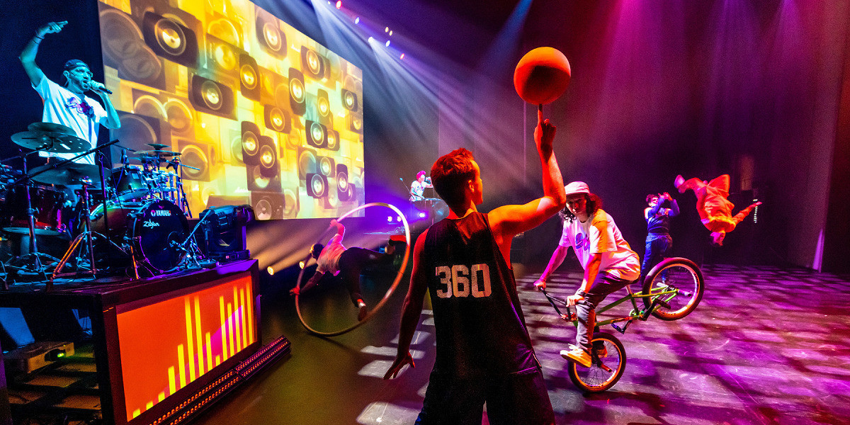 The 360 ALLSTARS stand in a group. There is a BMX rider, a basketballer, 2 breakdancers, and 2 musicians, under red and white lighting.
