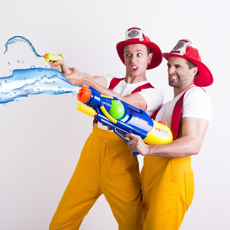 Two men dressed up as firefighters, older than they should be, playing with water pistols like it's very serious business