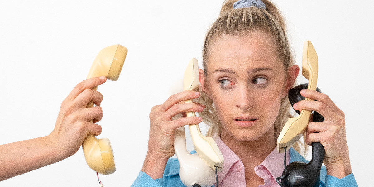 A phone is held up to a blonde woman who is also holding many phones, the woman looks at the incoming phone with concern.