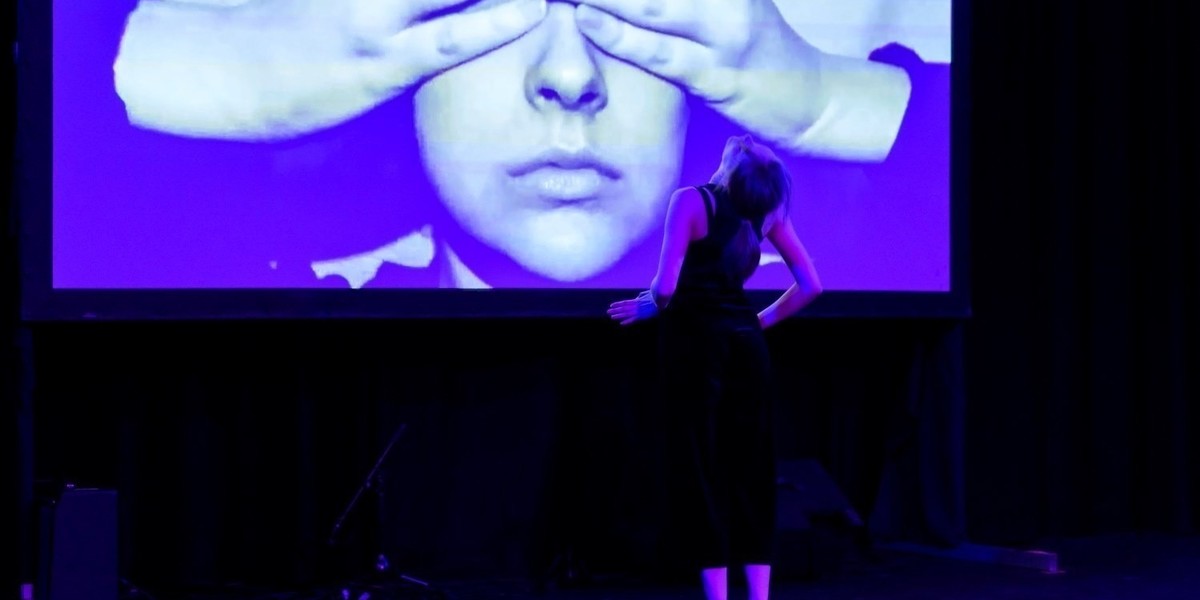 Amelia is standing facing away from the audience in front of the projection. She is in a slight back bend with her hands on her hips pointing towards the projection. The projection is a close up of Amelia's face and she has both hands covering her eyes. The lighting is a purple hue with a blue undertone.