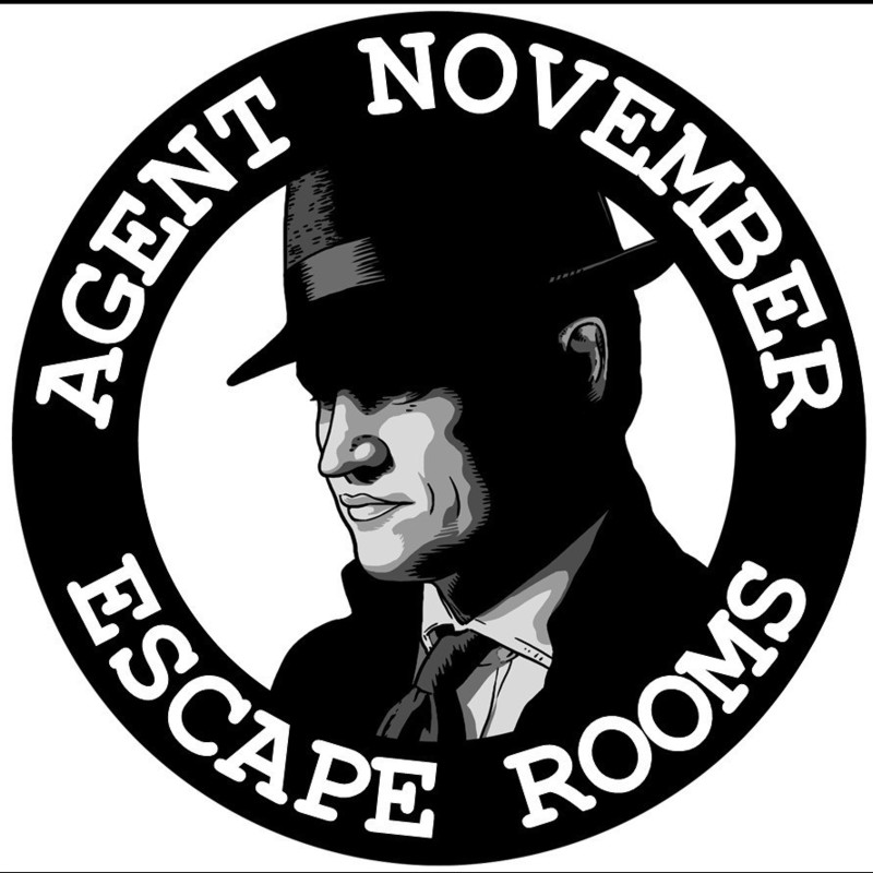 Agent November Escape Rooms: Crisis of Conscience - A thick black ring filled with white words saying "Agent November" across the top and "Escape Rooms" down the bottom. In the centre of the ring is a grayscale comic book style cartoon man wearing a black tie and a fedora had pulled down over his eyes.