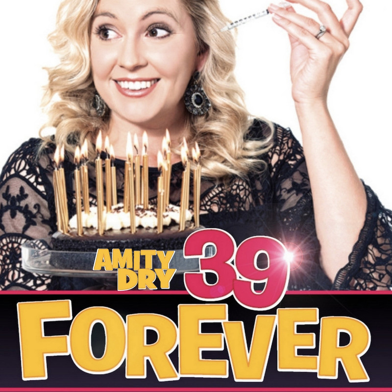 A photo of a woman with medium length blonde curly hair smiling and looking to the side. she is wearing a black lacy top and is holding a syringe in one hand. In front of her is a chocolate cake with multiple gold candles. The text below reads ‘Amity Dry 39 Forever’ in yellow and pink bold capital letters.