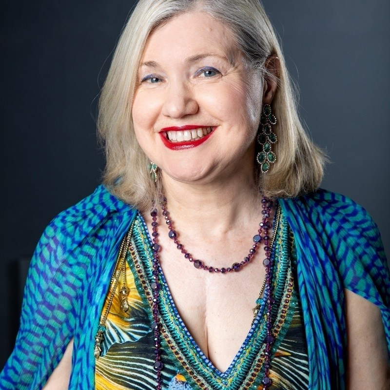 A headshot photo of a woman smiling. She has blonde hair that sits on her shoulders and is wearing red lipstick, large green gemstone earrings, purple necklace and a blue patterned dress.
