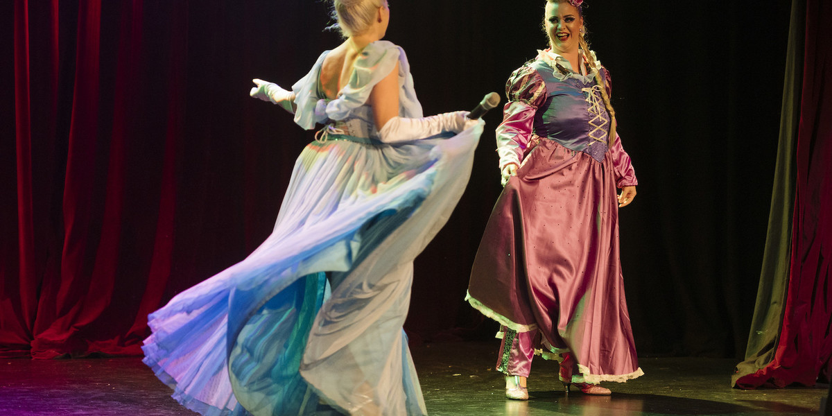 Performer dressed as Cinderella joyfully runs towards another,  performer, who is seated and dressed as Rupunzel from the Disney classics.