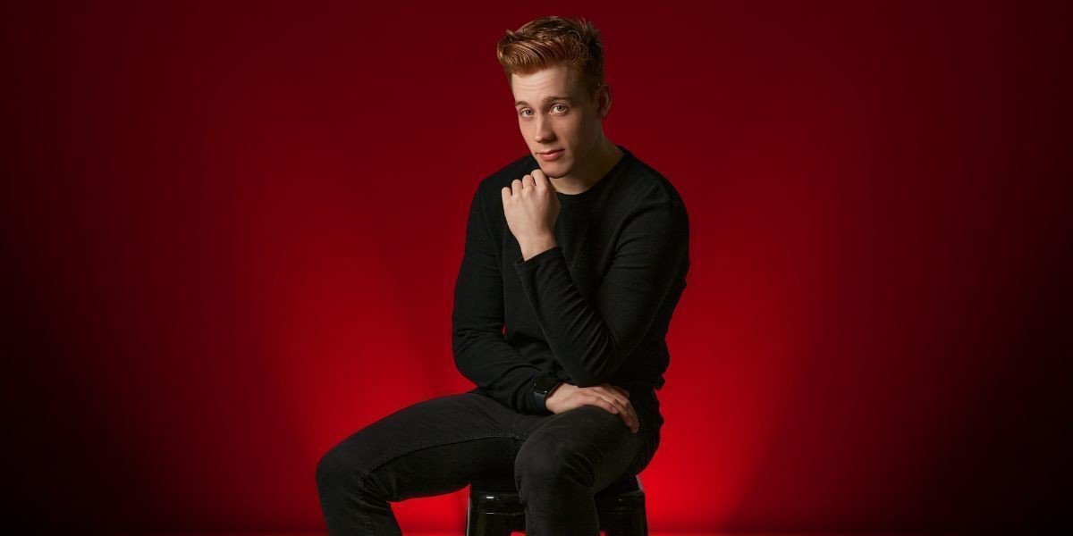 Daniel sitting on a stool with a smirk, his hand to his chin and a deep red background behind him.