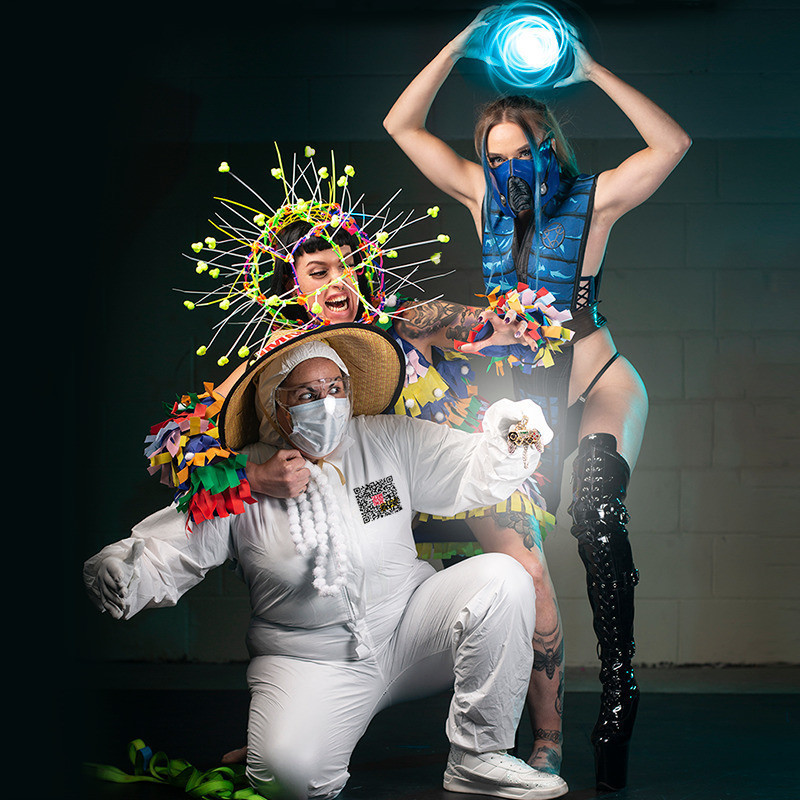 Vauxx dressed as a sexy Sub Zero wields a blue orb of light above her head. Saskia dressed as COVID stretches out from behind Mema Random dressed as a COVID Marshall version of Raiden holding a gold video game controller medallion.