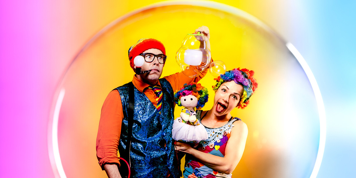 clown, puppet and man inside a bubble. The man holds a cube shaped bubble whilst blowing a white bubble out of his pipe