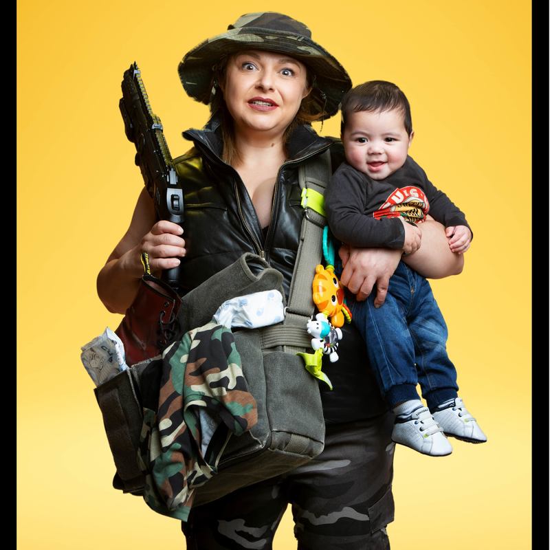 Vida Slayman in Comedy of Terrors - A woman dressed in military uniform looks perplexed as she carries a baby in one arm, a machine gun in the other, with an army bag full of military and baby stuff strapped across her chest.