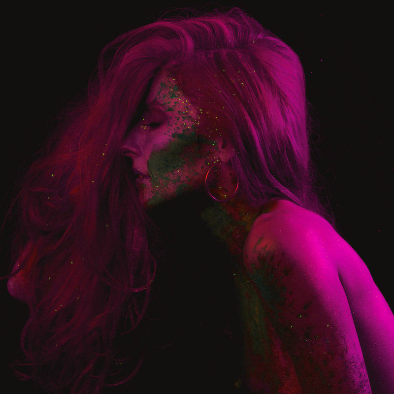 SOMNIA - A headshot image of a person facing the side with long hair covering their face. They have splotches of green paint over their face and body.