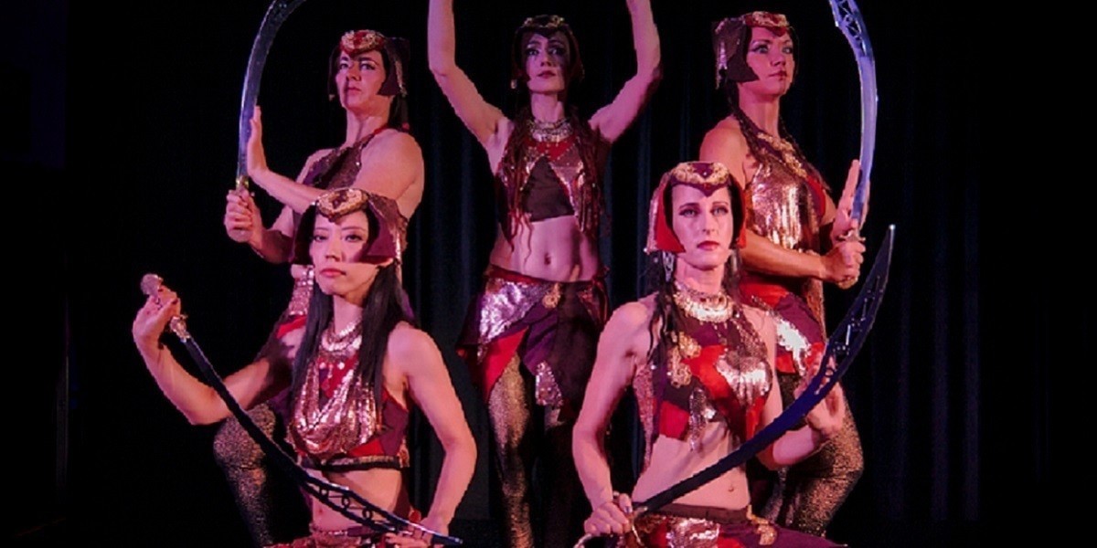 Five dancers, dressed in costumes resembling female warriors, shades of red with gold metallic fabric. Each dancer holds a sword, swords are held in a way that together they form a circle. Black background and warm lighting.