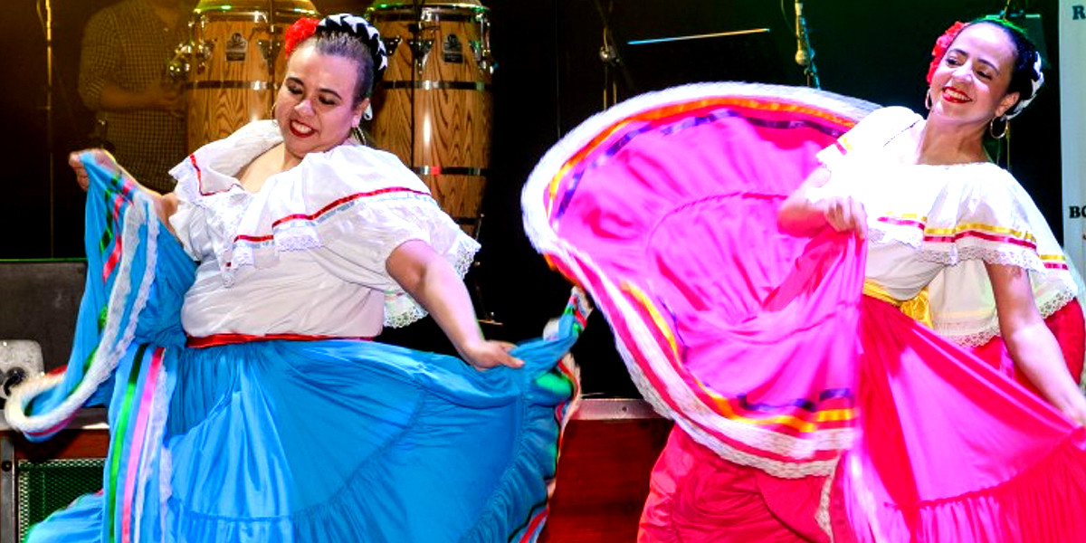 2 Mexican Revolution folk dancers in big swirly traditional Mexican long skirts. Dancer on the left is in a bright blue skirt and dancer on the right is dancing in a long hot pink skirt, they are both wearing while off the shoulder blouses