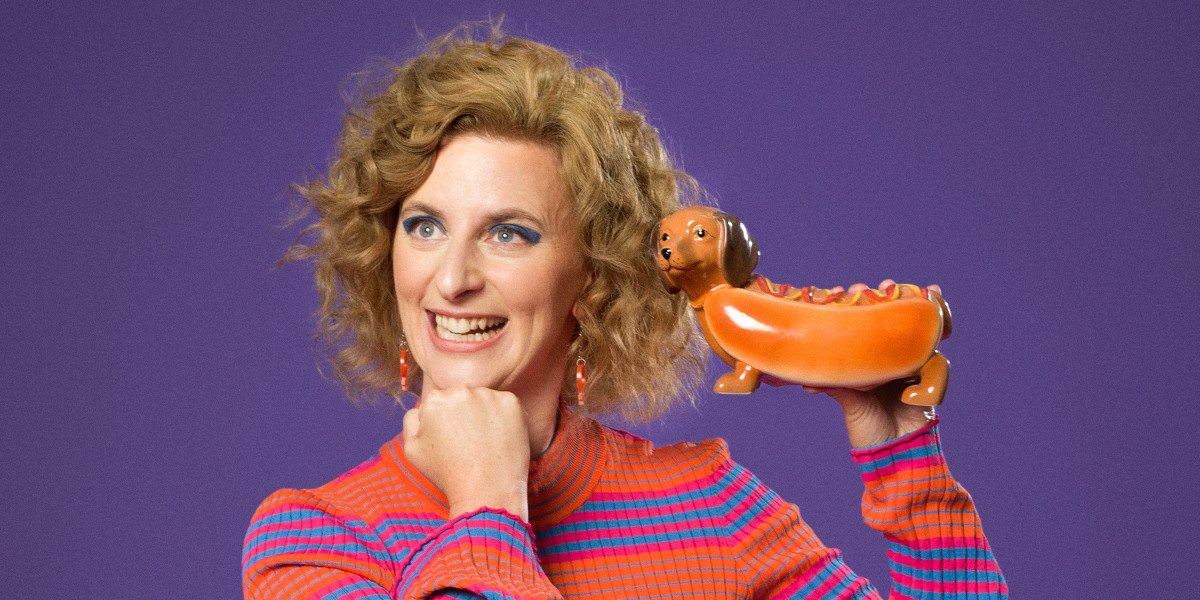 Felicity Ward - I'm Exhausting! - Felicity ward, wearing a bright orange, pink and blue striped top holding a ceramic "hot dog" Dachshund