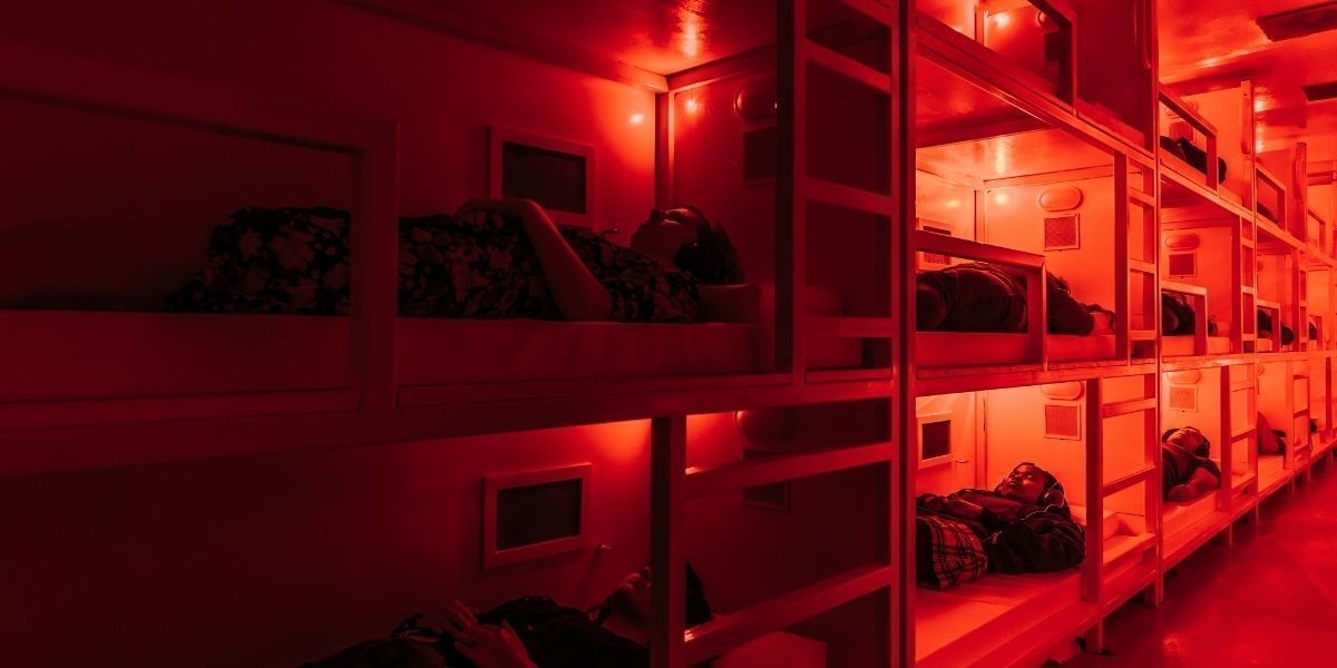 Interior of coma container, with people lying on bunk beds wearing headphones in a red light.