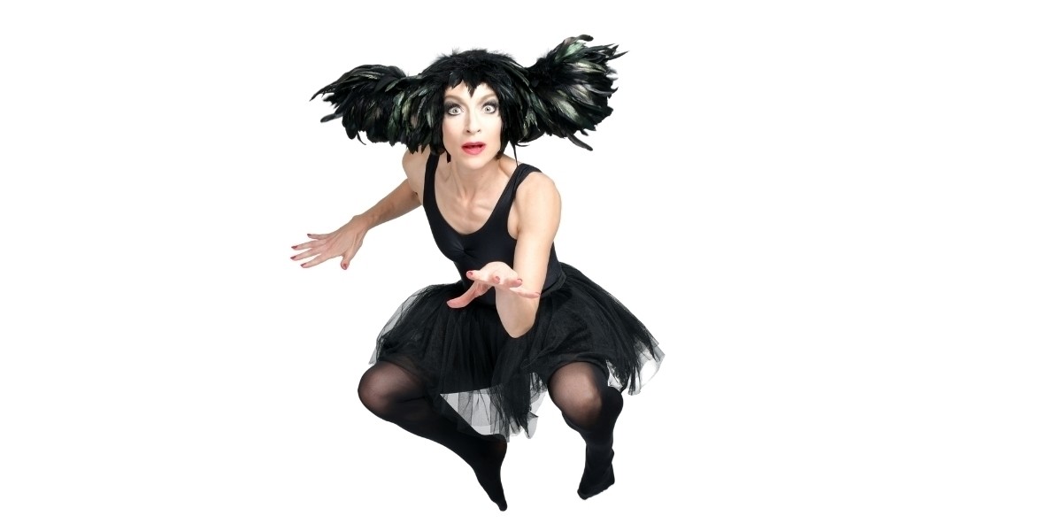 Sarah-Louise Young is suspended mid-jump, legs in a crouch position, jazzy arms. She wears a black feather headdress, teamed with a black tutu, leotard and dark black stockings with no shoes).