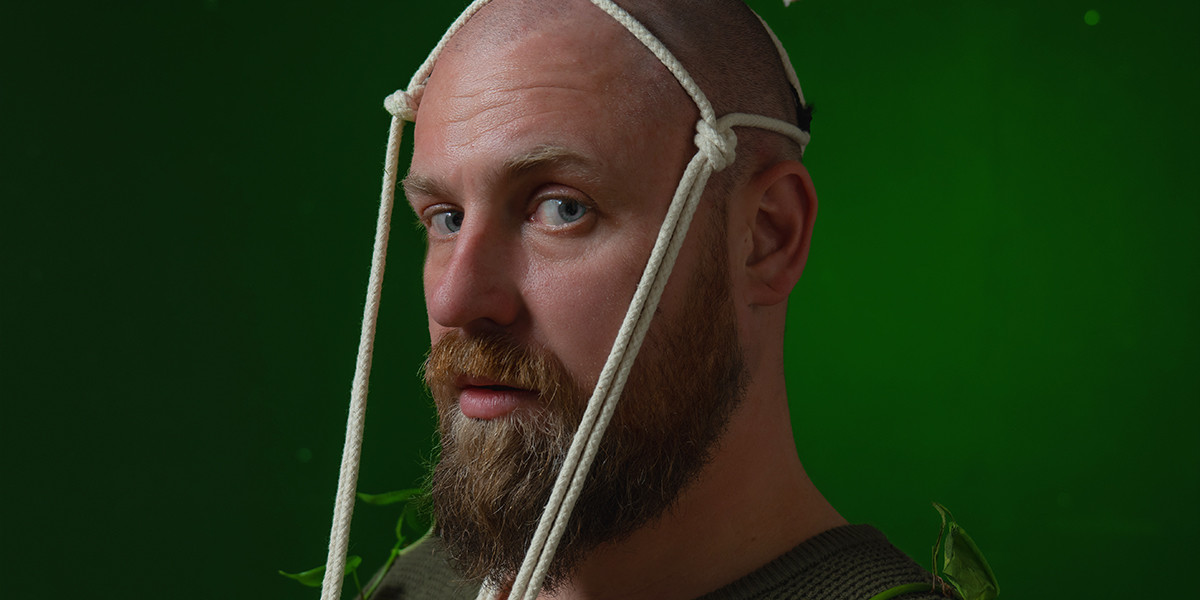 A man covered in vines wearing a macrame plant holder on his head looking directly into the lens.
